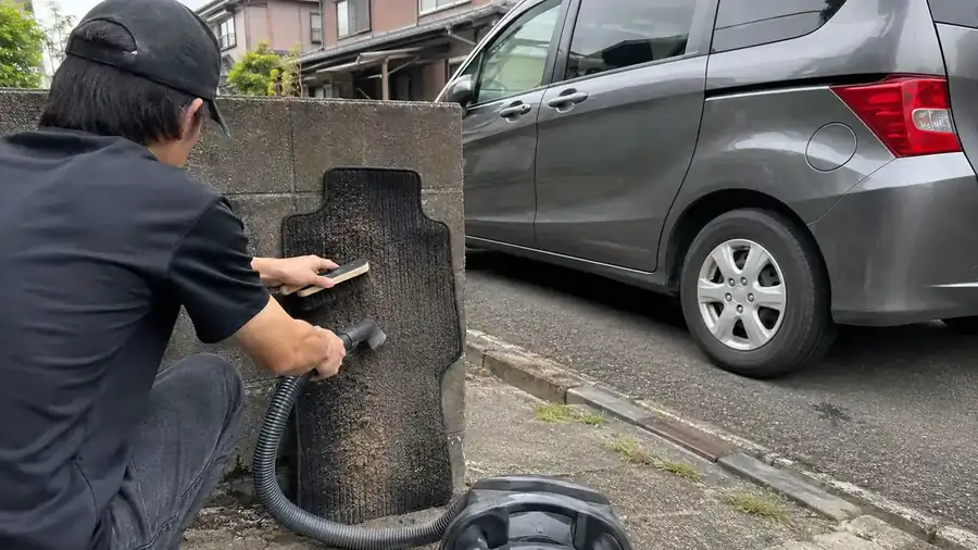 車から外したフロアマットを立てかけ、ブラシでこすりながら砂とホコリを掃除機で吸い取っているところ