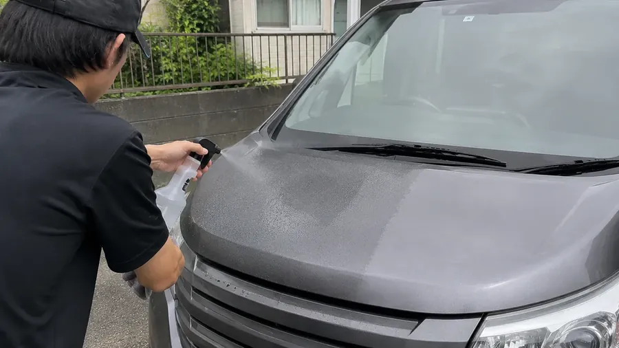 ミニバンのボンネットに、スプレーボトルで水なし洗車用のケミカルを吹き付けているところ。吹き付けた範囲だけ汚れが浮いて色が変わっている