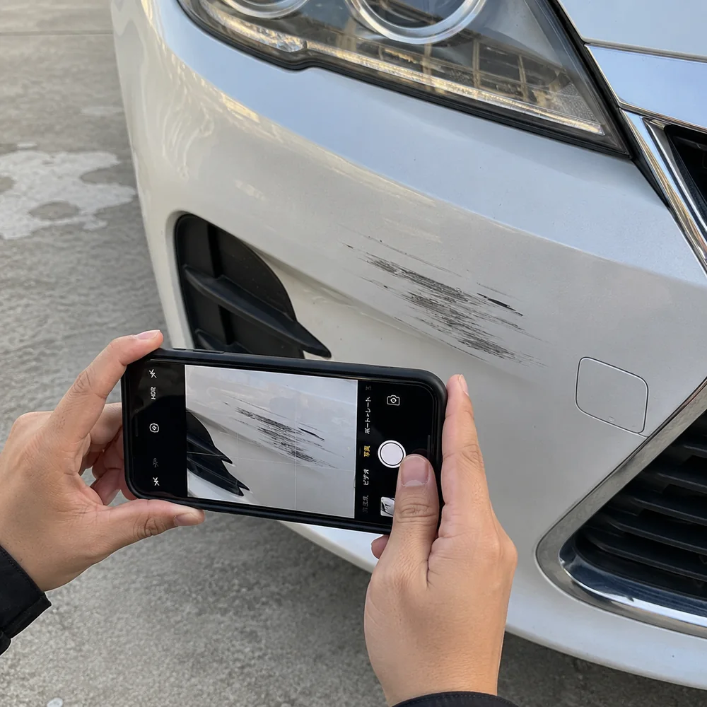 作業前の傷チェック記録。白い車のフロントバンパーにもとからある擦り傷を、スマートフォンで撮影しているところ