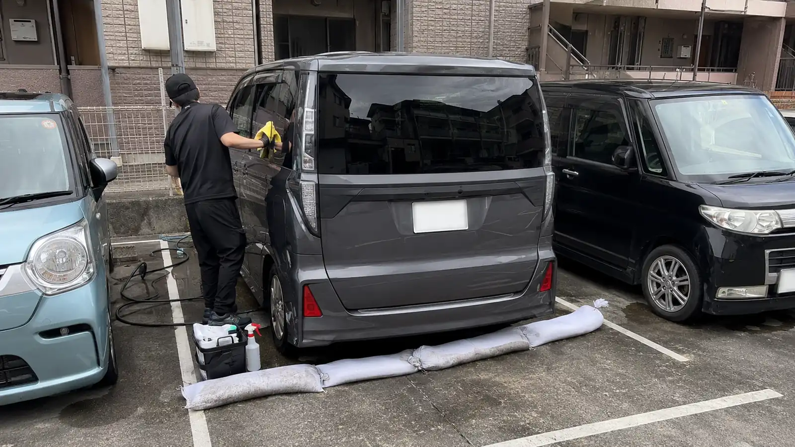 集合住宅の屋外駐車場で、両隣に車が停まったままの区画1台分を使ってグレーのミニバンを施工しているところ。車体の後ろには水が区画の外へ流れ出ないように白い吸水土のうを並べ、足元にケミカルを入れた道具箱を置いている
